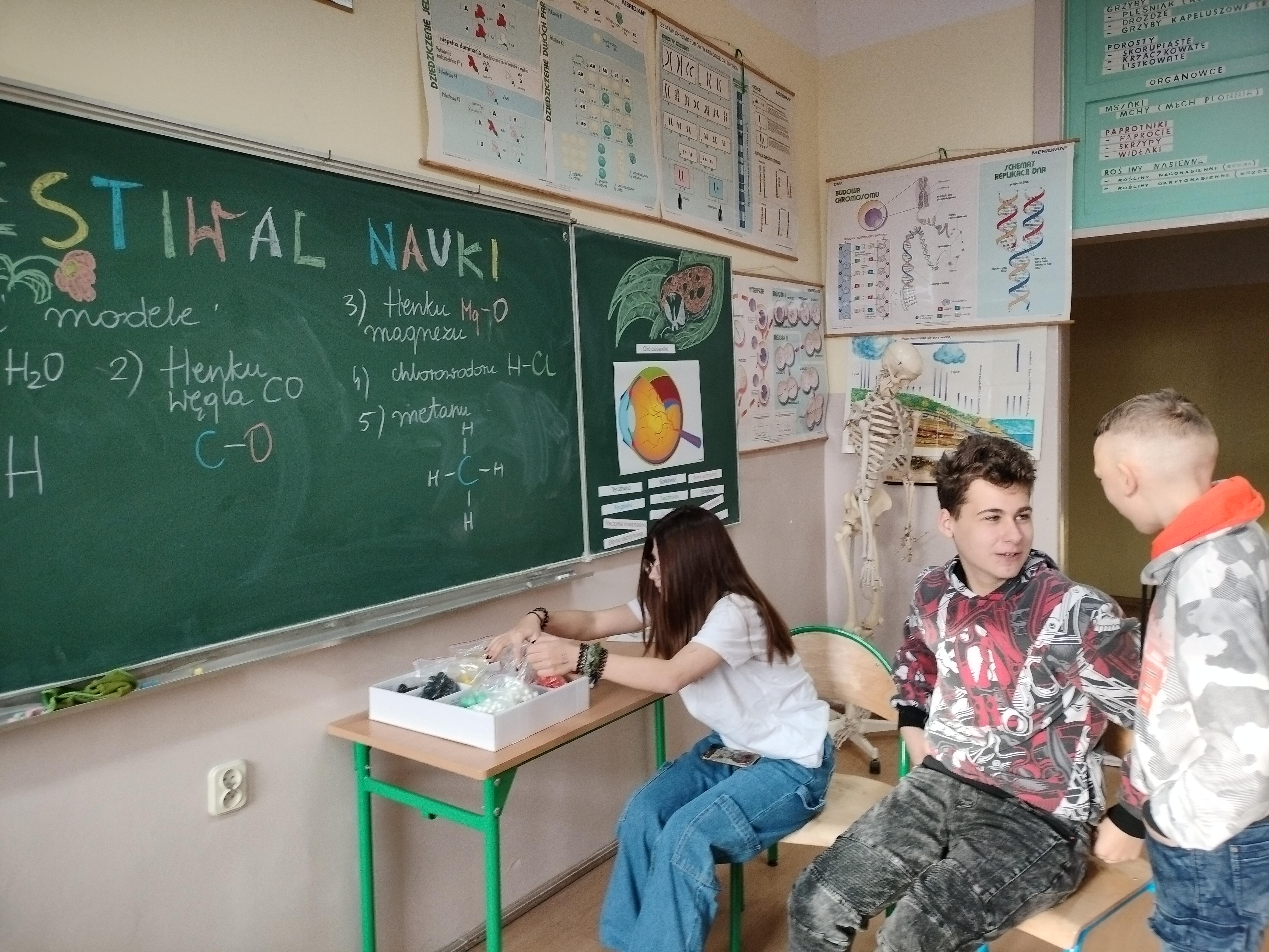 Po lewej stronie na scianie kolorowe napisy na tablicy i plansze. Po prawej troje osób
