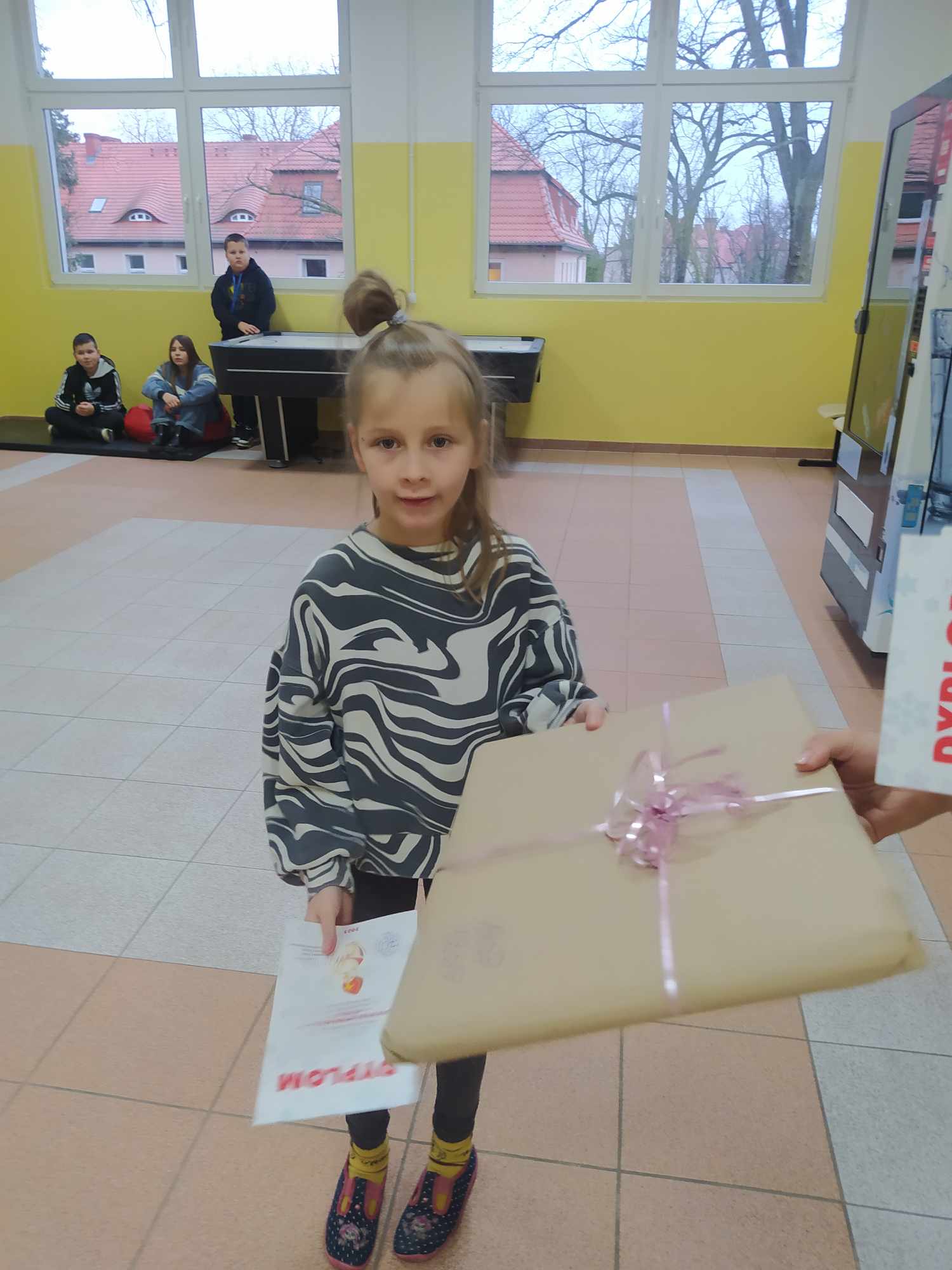 W pomieszczeniu stoi osoba. Z tylu przy oknie stoi grupa osób