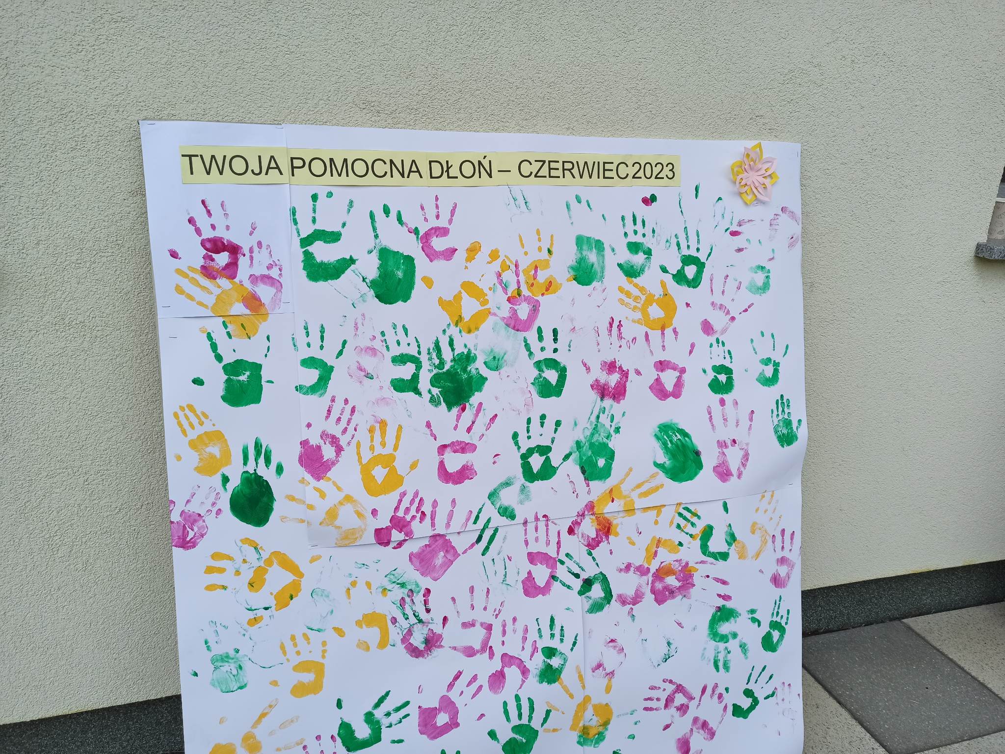 Na bialej tablicy kolorowe ślady dłoni. Na górze napis. 