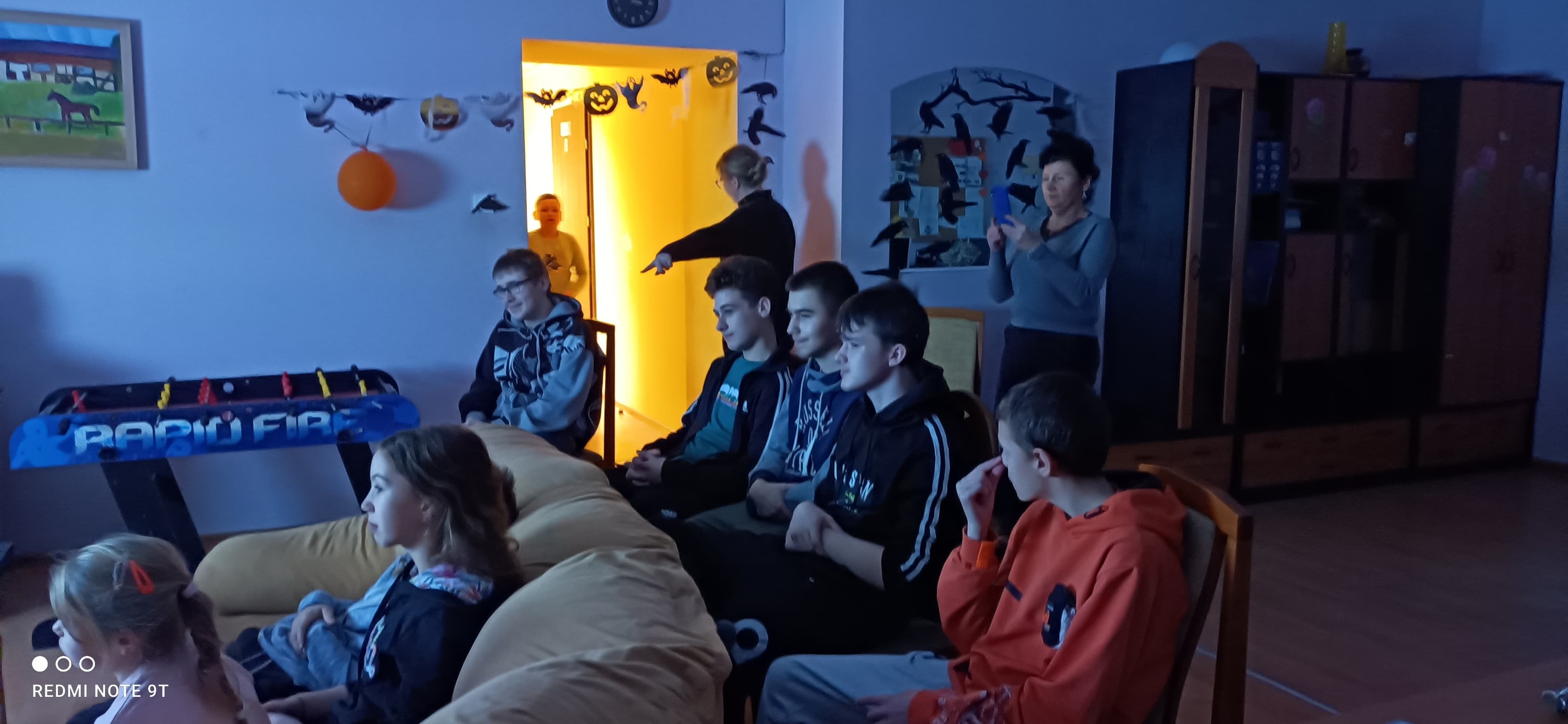 Na środku siedzi grupa uczniów, po prawej stoi postać