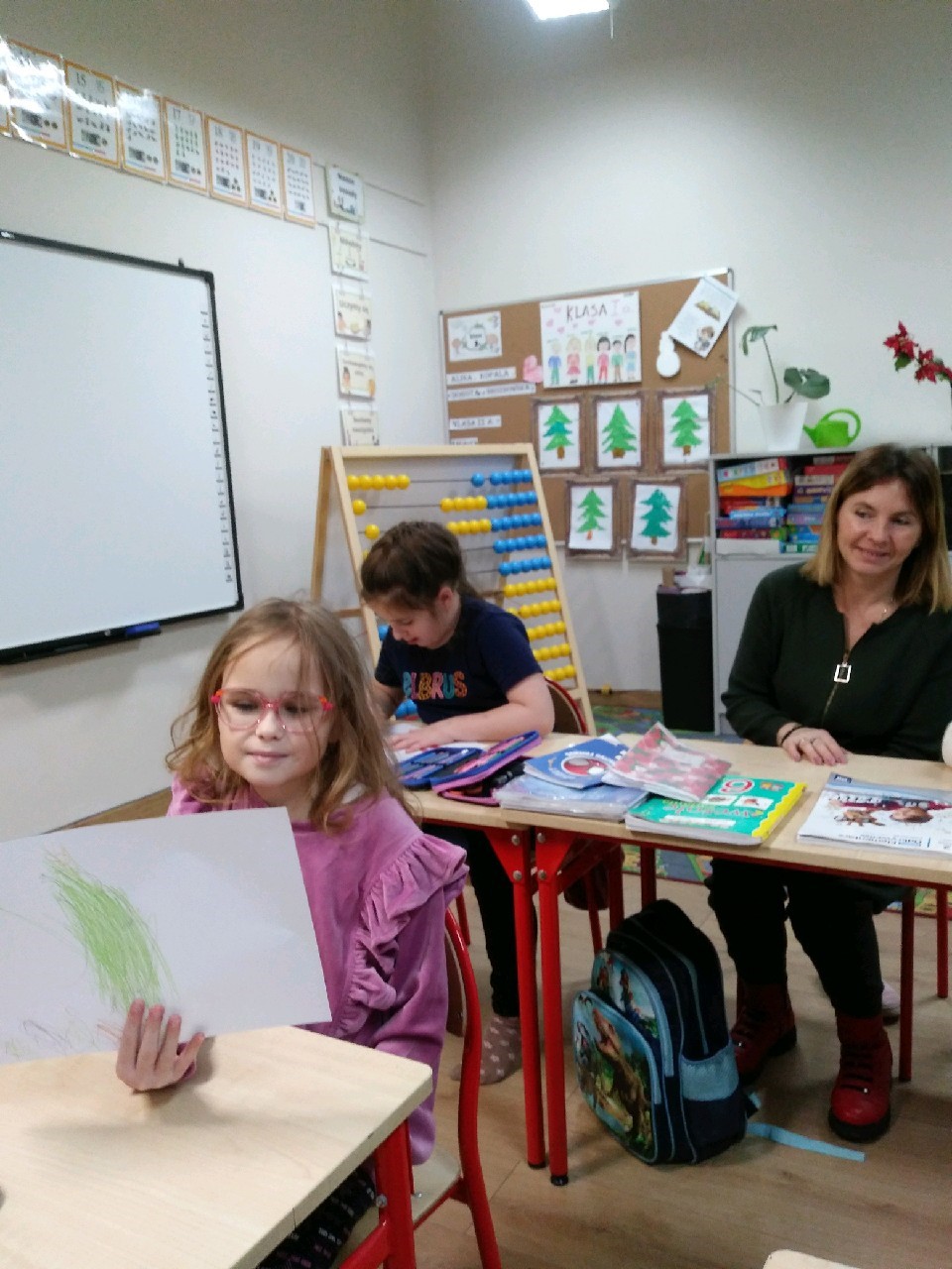 Na obrazku widać : trzy osoby, ławki, kartki, zeszyty